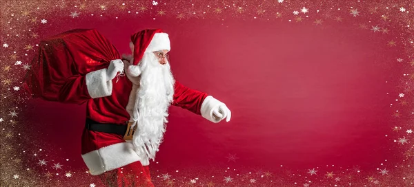 Πορτρέτο του crouching Santa Claus μεταφέρουν τεράστιο κόκκινο σάκο με δώρα σε κόκκινο φόντο στούντιο. Απομόνωση — Φωτογραφία Αρχείου