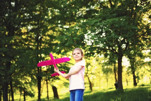 Niña lanza un avión de juguete al aire libre en el parque. Niño lanza un avión de juguete. Hermosa niña se para en la hierba y lanza un avión de juguete rosa — Foto de Stock