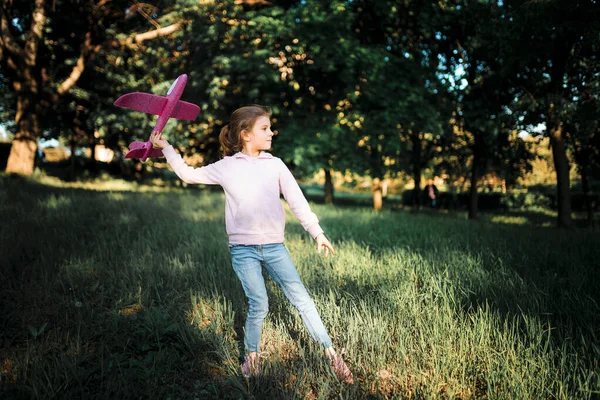 Το κοριτσάκι εκτοξεύει ένα αεροπλάνο-παιχνίδι στον αέρα στο υπαίθριο πάρκο. Το παιδί εκτοξεύει ένα παιχνίδι αεροπλάνο. Όμορφο κοριτσάκι στέκεται στο γρασίδι και εκτοξεύει ένα ροζ παιχνίδι αεροπλάνο — Φωτογραφία Αρχείου
