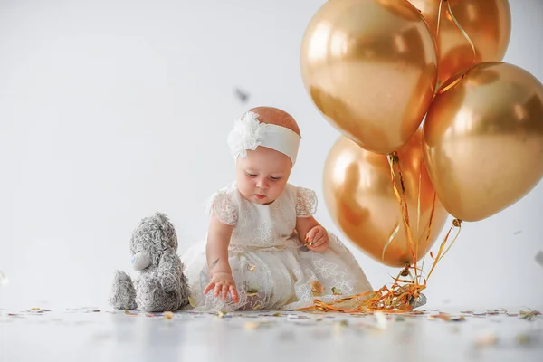 Uma menina de um ano de idade, sentada com um monte de balões dourados e taddy urso no fundo branco no estúdio — Fotografia de Stock
