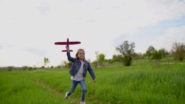 En liten flicka kastar ett leksaksplan i luften i parken utomhus. Barnet avfyrar ett leksaksplan. Vacker liten flicka springer på gräset och lanserar en rosa leksak plan — Stockvideo