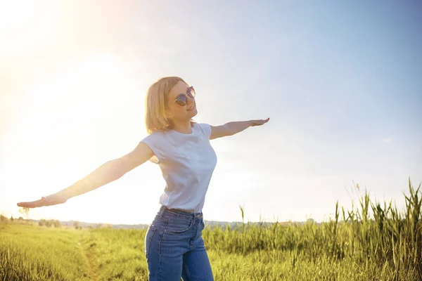 Egy nő fehér pólóban, napszemüvegben és farmerban széttárt karokkal utánozza a repülőt naplementekor. Nyár. Nyaralás légi utazás koncepciója — Stock Fotó