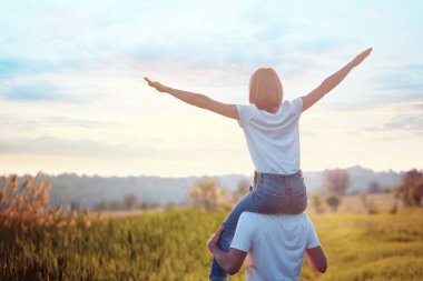 Genç adamın mutlu kız arkadaşını omuzlarında taşıyan portresi, uçağın taklidini yaparak kollarını ayırdı. Yaz. Tatil hava yolculuğu konsepti. Gün batımı
