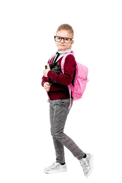 Child with schoolbag. Girl in white shirt and gray pants, rounded glasses, hold a book, school bag and posing like model. Isolated on white — ストック写真