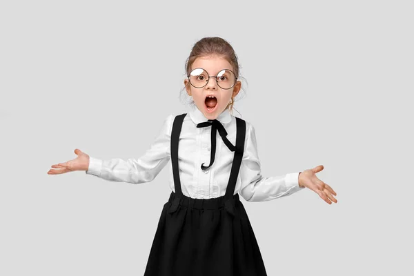 Εκπληκτικό κορίτσι με σχολική στολή, με στρογγυλά γυαλιά άπλωσε τα χέρια της στις πλευρές αγανακτισμένη. Στιγμιότυπο. Μεμονωμένο γκρι φόντο — Φωτογραφία Αρχείου