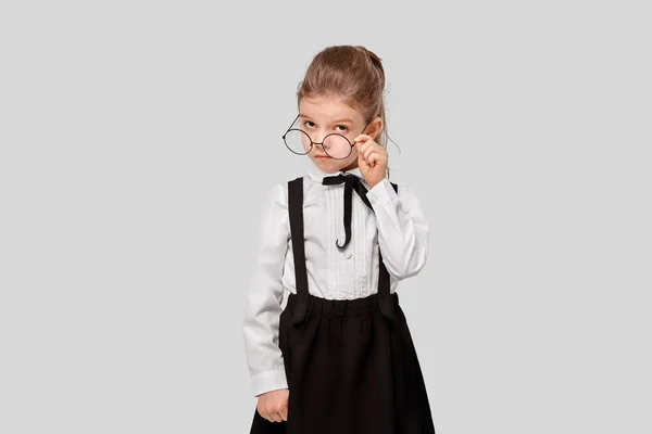 Girl in a school uniform holds round glasses and looks inquiringly at the camera. Studio shot. Isolated gray background — Stock Photo, Image