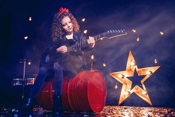 Portrait of a Beautiful rock girl with curly hair wearing leather jacket, boots playing the electric guitar while sitting on a red tank in recording studio