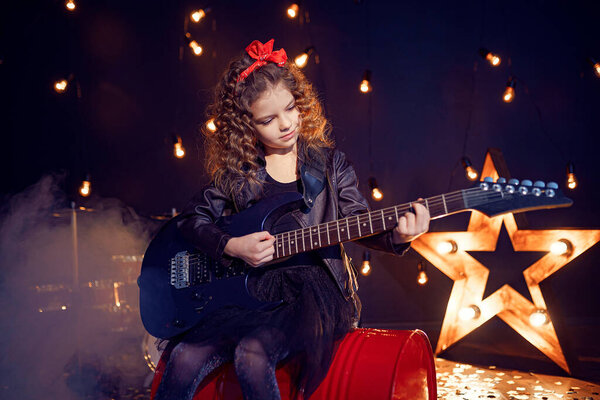 Portrait of a Beautiful rock girl with curly hair wearing leather jacket, boots playing the electric guitar while sitting on a red tank in recording studio