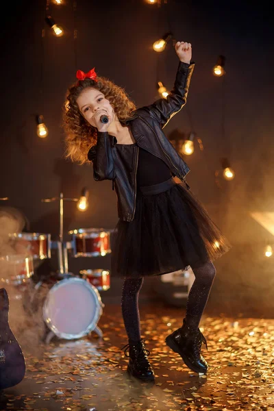 Gyönyörű lány göndör haj visel bőr kabát, csizma énekelni egy vezeték nélküli mikrofon karaoke, mint a rock sztár a stúdióban vagy a színpadon. Füst a háttérben — Stock Fotó