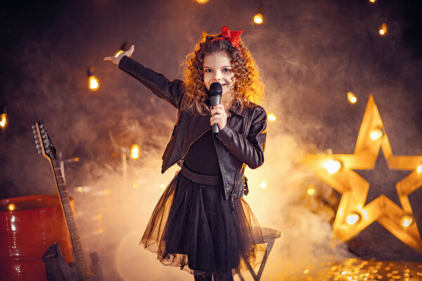 Beautiful girl with curly hair wearing leather jacket, boots sing into a wireless microphone for karaoke like rock star in recording studio or stage. Smoke on background