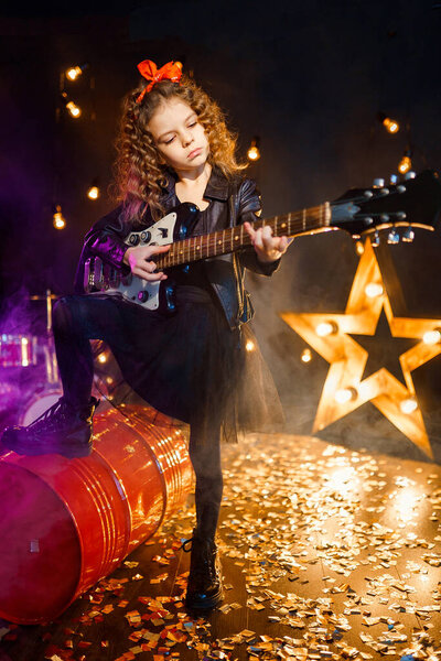 Portrait of a Beautiful rock girl with curly hair wearing leather jacket, boots playing the electric guitar behind a red tank in recording studio