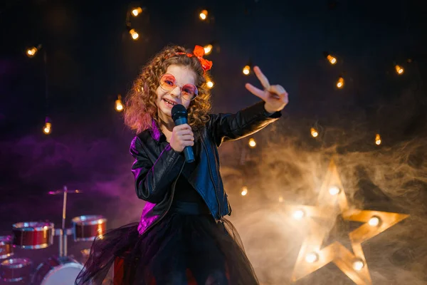 Menina bonita com cabelo encaracolado usando jaqueta de couro e óculos de sol vermelhos cantar em um microfone sem fio para karaoke em estúdio de gravação ou palco — Fotografia de Stock