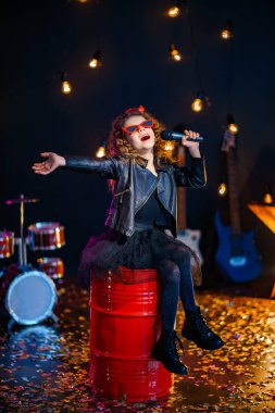 Beautiful girl with curly hair wearing leather jacket, red sunglasses sing into a wireless microphone for karaoke while sitting on red tank in recording studio or stage