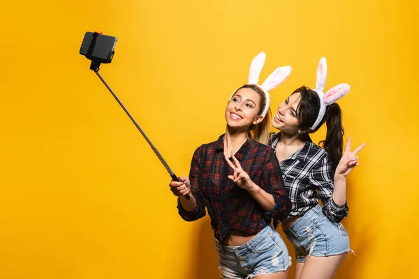 Dos jóvenes sexy mujer caucásica usando lindas orejas de conejo de Pascua haciendo selfie usando palo sobre fondo aislado amarillo — Foto de Stock