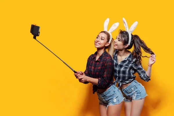 Two Young sexy caucasian woman wearing cute easter rabbit ears making selfie using stick over yellow isolated background — Stockfoto