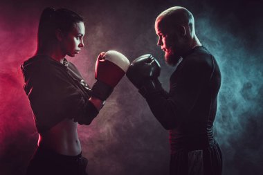 Boks ve savunma dersinde antrenörle egzersiz yapan bir kadın. Stüdyo, arka planda sigara içmek. Birbirinize agresif bir şekilde bakın. Önde dur.