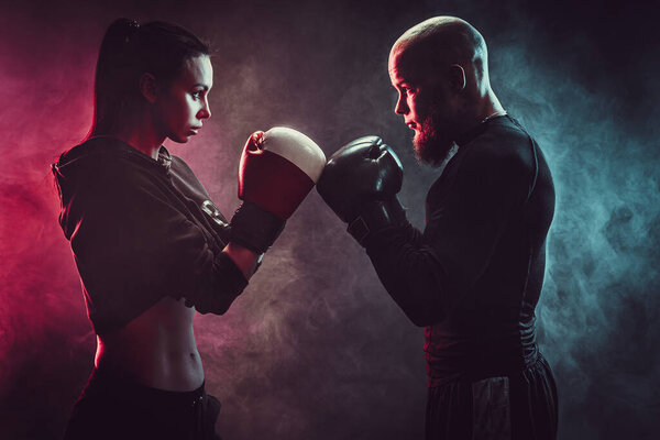 Woman exercising with trainer at boxing and self defense lesson, studio, smoke on background. Aggresively look each other. Stand in front