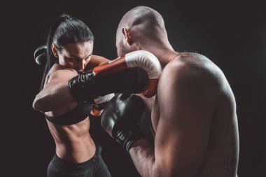 Gömleksiz kadın boks antrenörüyle egzersiz yapıyor ve savunma dersleri alıyor, stüdyo, arka planda sigara içiyor. Kadın ve erkek kavga eder.