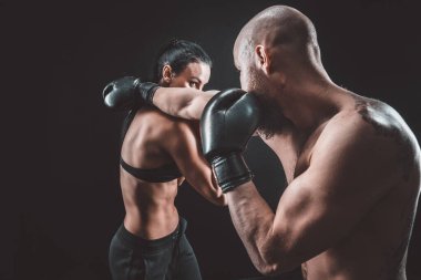 Gömleksiz kadın boks antrenörüyle egzersiz yapıyor ve savunma dersleri alıyor, stüdyo, arka planda sigara içiyor. Kadın ve erkek kavga eder.