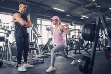 Odaklanmış ve motive olmuş spor kıyafetli genç kız bacak egzersizi yaparken yakışıklı kaslı antrenör onu izliyor. .