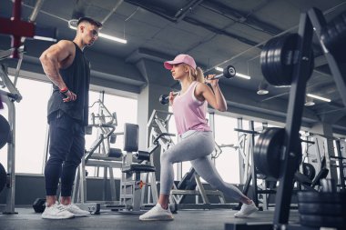 Odaklanmış ve motive olmuş spor kıyafetli genç kız bacak egzersizi yaparken yakışıklı kaslı antrenör onu izliyor. .