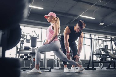 Odaklanmış ve motive olmuş spor kıyafetli genç kız bacak egzersizi yaparken yakışıklı kaslı antrenör onu izliyor. .