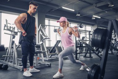 Odaklanmış ve motive olmuş spor kıyafetli genç kız bacak egzersizi yaparken yakışıklı kaslı antrenör onu izliyor. .