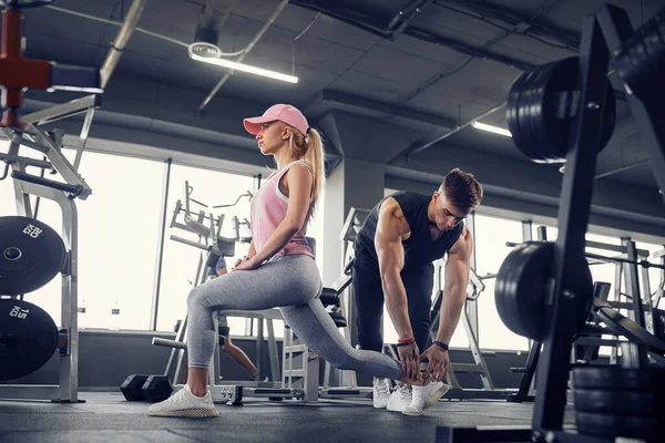 Odaklanmış ve motive olmuş spor kıyafetli genç kız bacak egzersizi yaparken yakışıklı kaslı antrenör onu izliyor. .