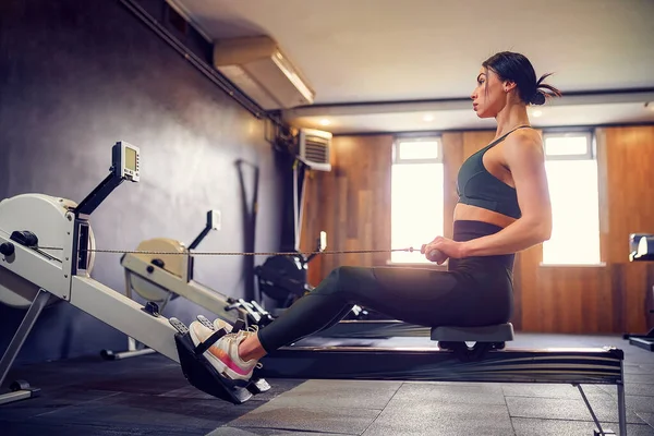 Mujer joven decidida haciendo ejercicio en la máquina de fila en el gimnasio —  Fotos de Stock