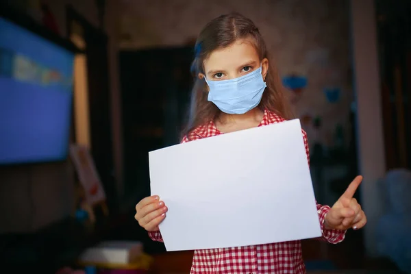 Liten flicka i en skyddande mask, pyjamas håller ett blankt papper med ena handen och den andra pekar finger när du står i rummet hemma, Skydd mot coronavirus — Stockfoto