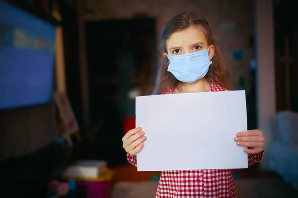 Liten flicka i en skyddande mask, pyjamas håller ett tomt papper när du står i rummet hemma, Skydd mot coronavirus — Stockfoto
