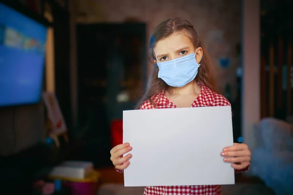 Liten flicka i en skyddande mask, pyjamas håller ett tomt papper när du står i rummet hemma, Skydd mot coronavirus — Stockfoto