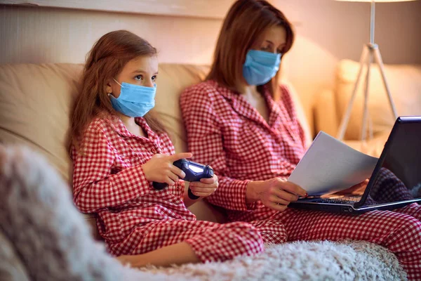 Kvinna i pyjamas med anteckningsbok och papper som arbetar hemifrån bär skyddsmask medan hennes barn, dotter spelar datorkonsol spel — Stockfoto