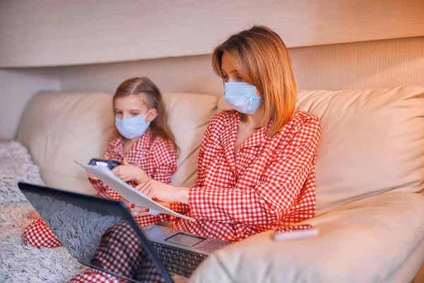 Kvinna i pyjamas med anteckningsbok och papper som arbetar hemifrån bär skyddsmask medan hennes barn, dotter spelar datorkonsol spel — Stockfoto