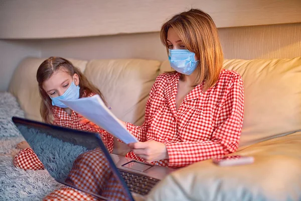 Kvinna i pyjamas med anteckningsbok och papper som arbetar hemifrån bär skyddsmask medan hennes barn, dotter spelar datorkonsol spel — Stockfoto