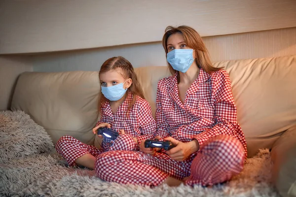 Kadın ve genç kız pijama giyiyor ve tıbbi koruyucu maskeler takıyor oturma odasındaki koltukta video oyunu kontrolörleriyle ev izolasyonu otomobil karantinası, covid-19