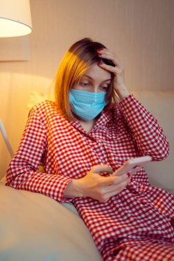 Cerrahi maske takan genç Avrupalı kadın Coronavirus, covid-19 salgını sırasında evde kalıyor ve akıllı telefondan haber veya uygulama izliyor