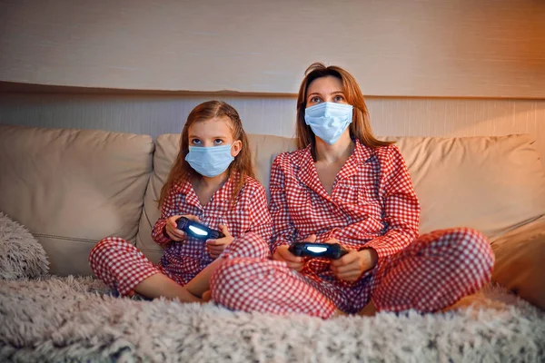 Woman and young girl wearing pajamas and medical protective masks sitting on sofa in living room with video game controllers at home isolation auto quarantine, covid-19