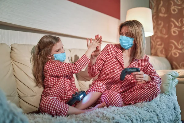 Kvinna och ung flicka bär pyjamas och medicinska skyddsmasker sitter på soffan i vardagsrummet med videospel styrenheter hemma isolering auto karantän, covid-19 — Stockfoto