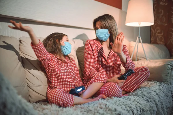 Kvinna och ung flicka bär pyjamas och medicinska skyddsmasker sitter på soffan i vardagsrummet med videospel styrenheter hemma isolering auto karantän, covid-19 — Stockfoto