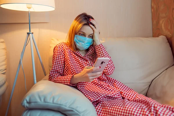 Ung europeisk kvinna bär kirurgisk ansiktsmask stanna hemma under coronavirus, covid-19 utbrott och ser nyheter eller ansökan på smartphone — Stockfoto