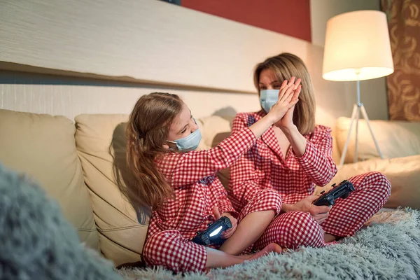 Woman and young girl wearing pajamas and medical protective masks sitting on sofa in living room with video game controllers at home isolation auto quarantine, covid-19