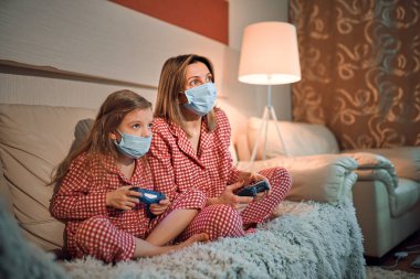 Kadın ve genç kız pijama giyiyor ve tıbbi koruyucu maskeler takıyor oturma odasındaki koltukta video oyunu kontrolörleriyle ev izolasyonu otomobil karantinası, covid-19