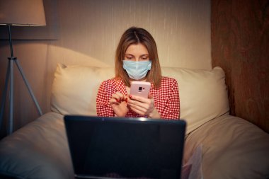 Cerrahi maske takan genç Avrupalı kadın Coronavirus, covid-19 salgını sırasında evde kalıyor ve akıllı telefondan haber veya uygulama izliyor