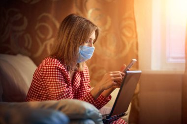 Cerrahi maske takan genç Avrupalı kadın Coronavirus, covid-19 salgını sırasında evde kalıyor ve akıllı telefondan haber veya uygulama izliyor