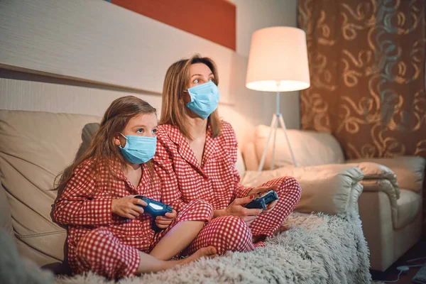 Kadın ve genç kız pijama giyiyor ve tıbbi koruyucu maskeler takıyor oturma odasındaki koltukta video oyunu kontrolörleriyle ev izolasyonu otomobil karantinası, covid-19