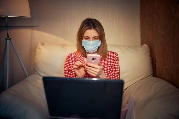 Jeune femme européenne portant un masque chirurgical rester à la maison pendant l'épidémie de coronavirus, covid-19 et regarder les nouvelles ou l'application sur smartphone — Photo