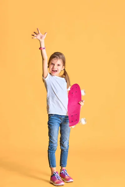 Elegante chica divertida con camiseta blanca, vaqueros azules y zapatillas de deporte, la celebración de monopatín sobre fondo amarillo — Foto de Stock