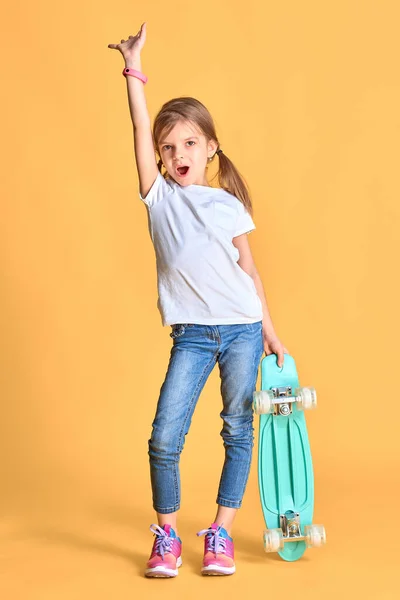 Elegante chica divertida con camiseta blanca, vaqueros azules y zapatillas de deporte, la celebración de monopatín sobre fondo amarillo — Foto de Stock