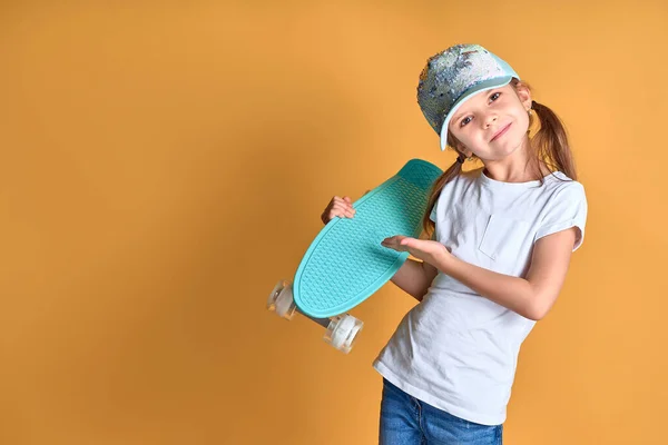 Κομψό κορίτσι φορώντας λευκό t-shirt, μπλε τζιν και πράσινο καπέλο, κρατώντας skateboard πάνω από κίτρινο φόντο — Φωτογραφία Αρχείου
